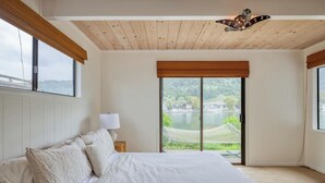 A bedroom with a large bed, a nightstand with a lamp, and a view of a lake through a sliding door. - Cozy 3BR by the lagoon w/ hot tub (Stinson Beach)