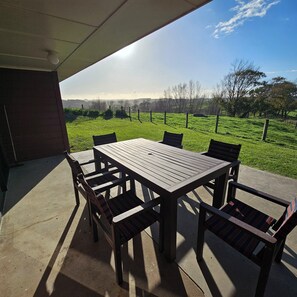Une terrasse extérieure avec une table et des chaises en bois, un sol en béton et une vue sur un champ verdoyant et des arbres.