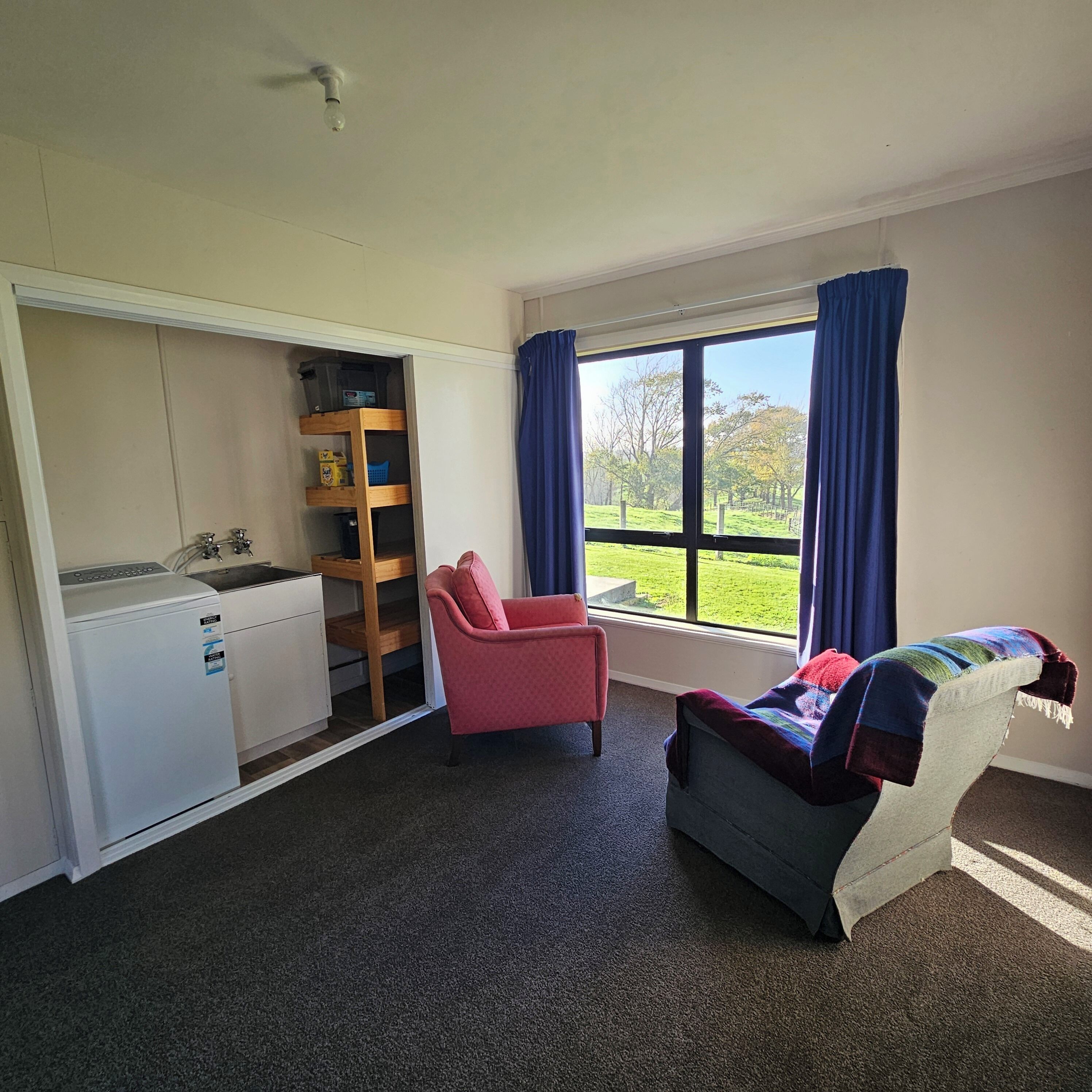 A room with a bed, a chair, a desk with a washing machine, a window with curtains, and a view of a green landscape.