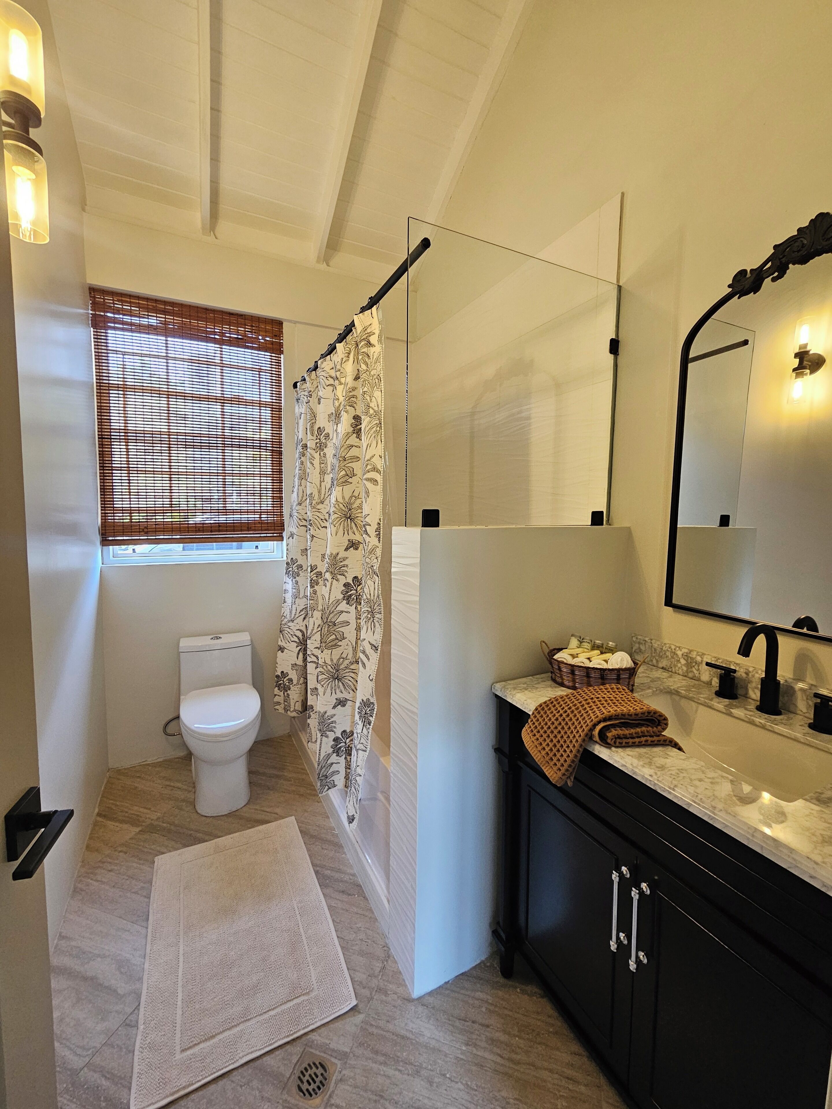 A bathroom with a toilet, shower curtain, sink, mirror, and a window with blinds.