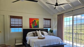 A bedroom with a large bed, two ceiling fans, a window with blinds, a painting, and a view of the outdoors.