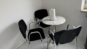 Una mesa pequeña y redonda con superficie de mármol, rodeada por cuatro sillas negras de estructura metálica. Un dispensador de toallas de papel blancas se encuentra sobre la mesa.