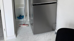 Una cocina compacta con un refrigerador de acero inoxidable, un estante blanco y una escoba en el suelo.