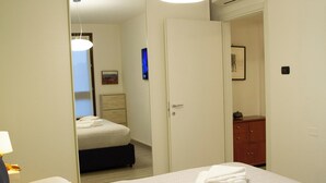 Habitación de hotel con cama, mesita de noche, lámpara, armario y puerta que da a otra habitación.