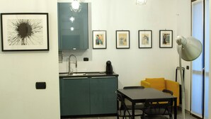 Une cuisine de taille réduite avec des meubles de couleur turquoise, une table à manger noire avec quatre chaises et un fauteuil jaune. Quatre œuvres d’art encadrées ornent le mur, et une lampe sur pied se trouve près de la fenêtre.