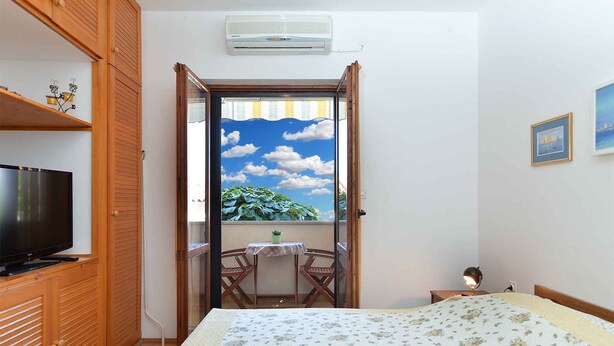 A bedroom with a bed, a television, a balcony with a table and chairs, and a view of the sky.