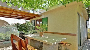 A patio area with a wooden bench and a table, surrounded by plants and a pergola.