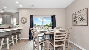 A modern kitchen with a dining area, featuring a large window, a clock on the wall, and a painting above the table.