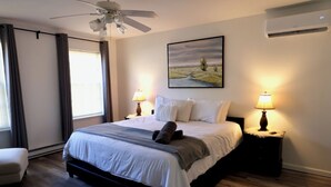 A bedroom with a bed, two bedside lamps, a ceiling fan, a window with curtains, and a painting on the wall.