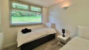 Un dormitorio con cama, mesita de noche con lámpara, ventana con persianas y vistas a zonas verdes.