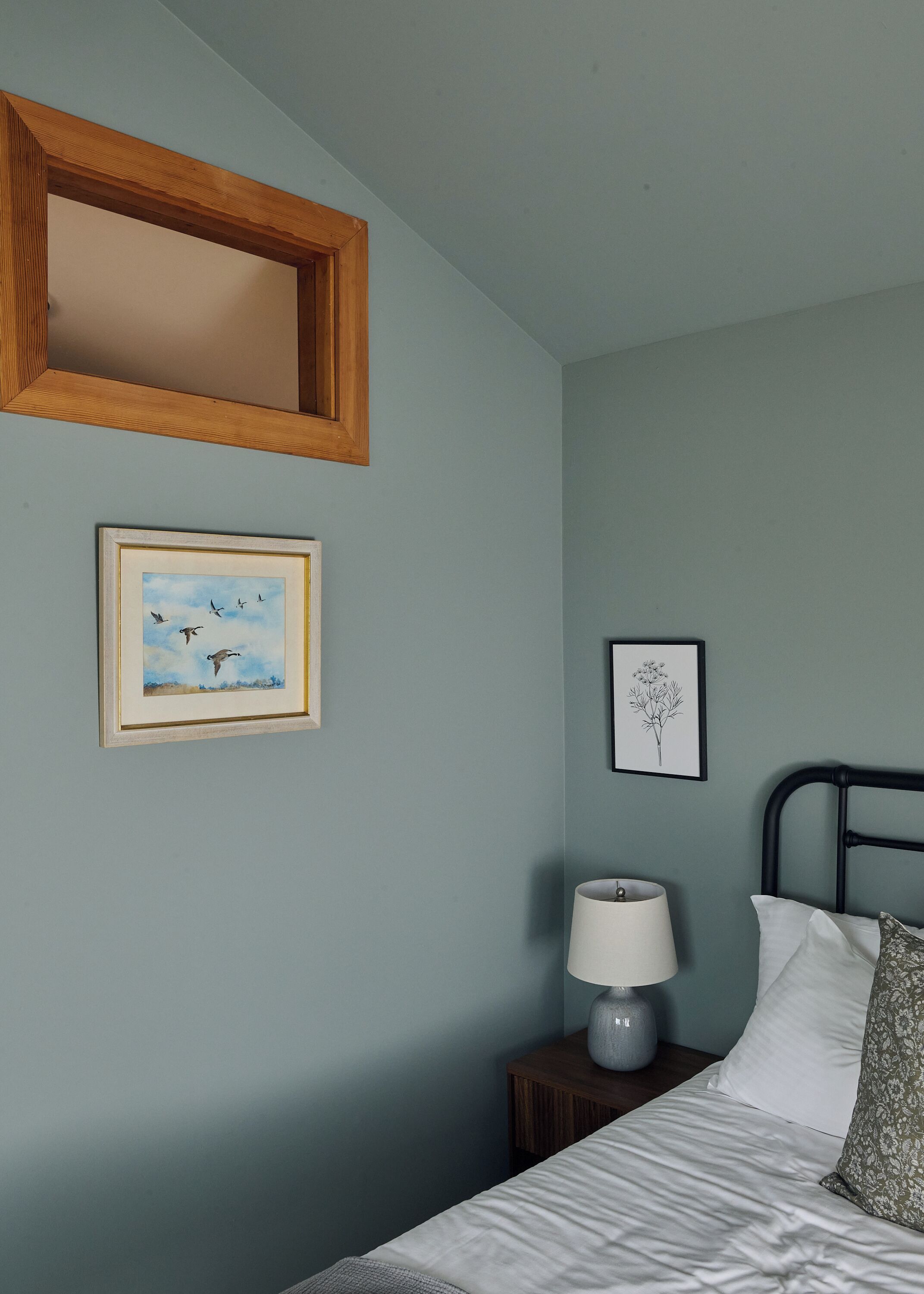 A bedroom with a bed, a nightstand, a lamp, a framed picture of birds, a framed botanical illustration, and a wooden-framed mirror.
