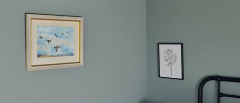 A bedroom with a bed, a nightstand, a lamp, a framed picture of birds, a framed botanical illustration, and a wooden-framed mirror.