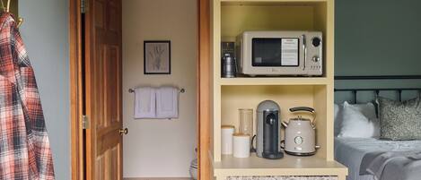 A bedroom with a bed, a bedside table with a kettle, a microwave, and a shelf with glasses and a basket.