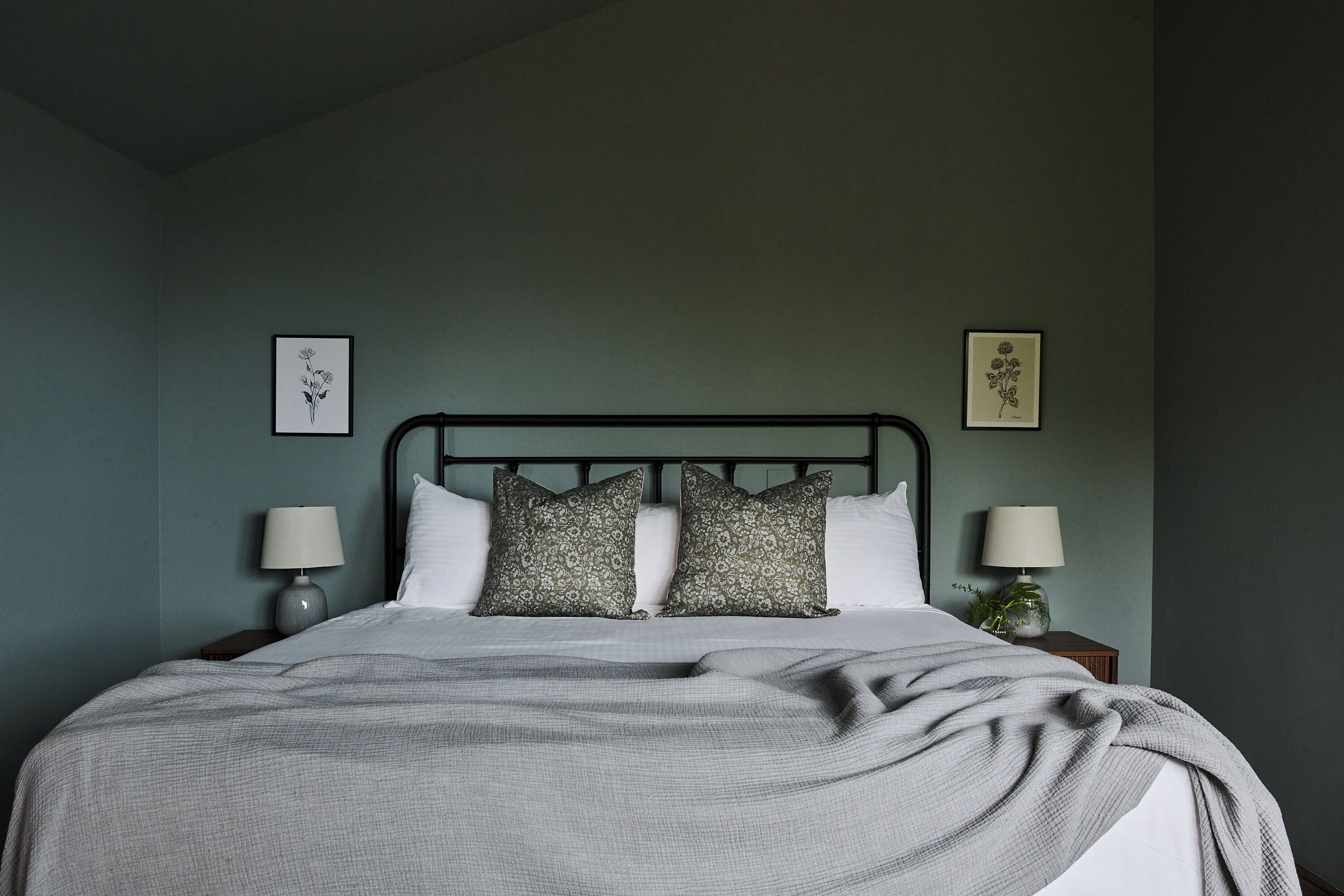 A neatly made bed with a grey blanket, two pillows, and a headboard. There are two bedside tables with lamps and framed pictures on the wall.