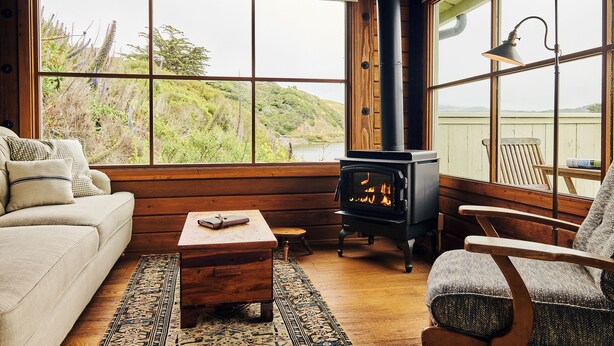 A cozy cabin interior with a large window, a wood-burning stove, a sofa, a chair, and a small table.