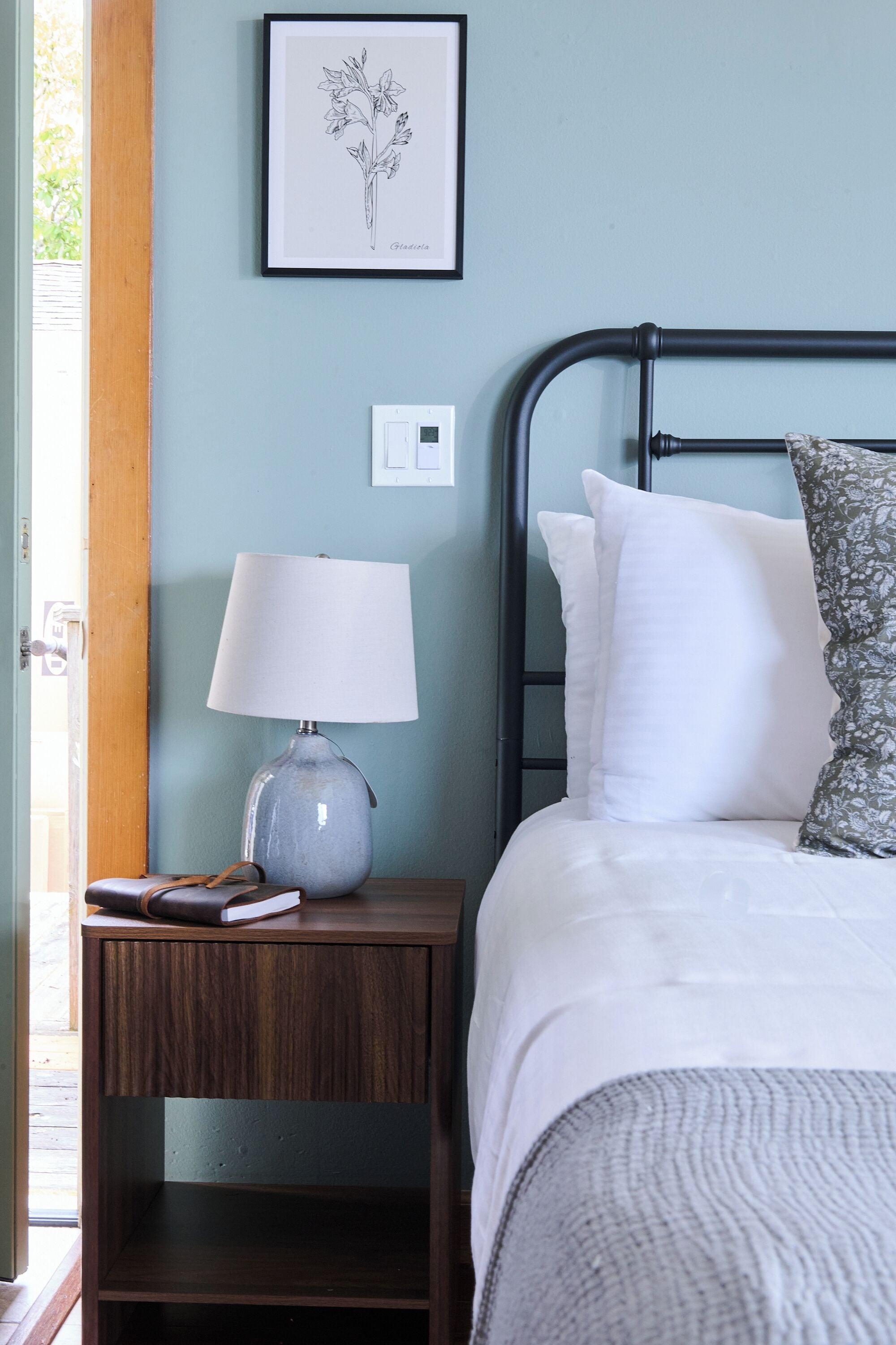A neatly made bed with white and patterned pillows, a bedside lamp, and a framed botanical illustration on the wall.