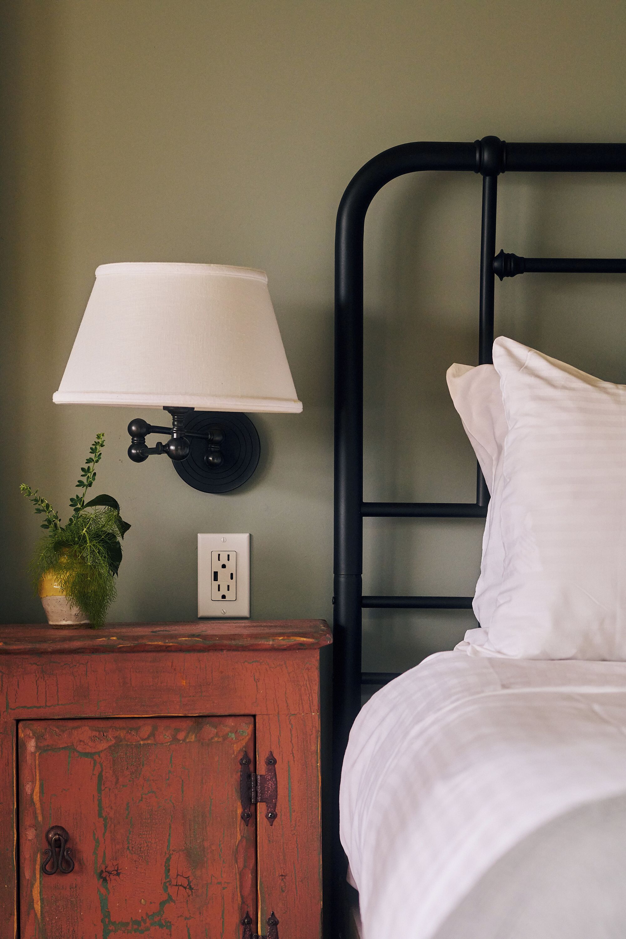 A bed with white bedding, a black metal headboard, a wall-mounted lamp, a small wooden nightstand with a potted plant, and a power outlet.