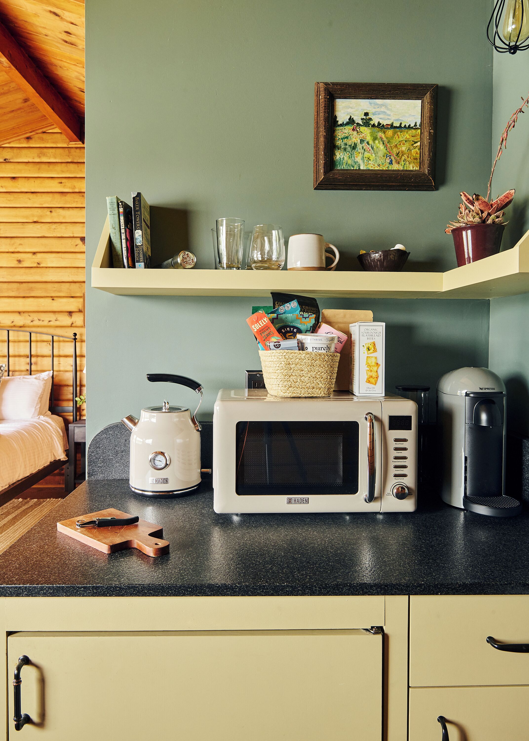 Een keuken met een magnetron, broodrooster en koffiezetapparaat. Op het aanrecht staat een mandje en daarboven een plank met boeken en glazen.