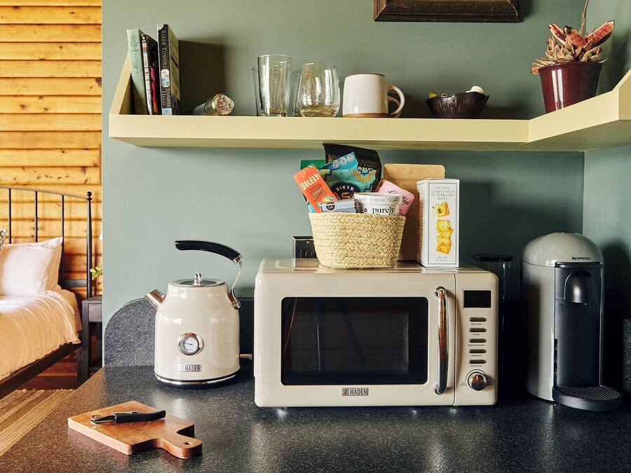 Een keuken met een magnetron, broodrooster en koffiezetapparaat. Op het aanrecht staat een mandje en daarboven een plank met boeken en glazen.