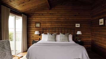 A wooden cabin bedroom with a bed, two nightstands, a chair, and a large window with curtains.