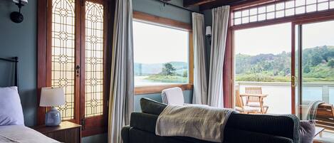 A bedroom with a bed, a nightstand, a lamp, a window with curtains, and a view of a lake and hills.
