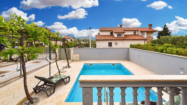 Ein rechteckiger Swimmingpool mit Fliesenverkleidung, umgeben von einer niedrigen Mauer und einem Balkongeländer.