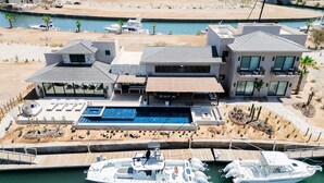 Una casa moderna de varios pisos con una gran piscina, barcos amarrados y un puerto deportivo.