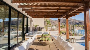 Un patio al aire libre con asientos blancos acolchados, macetas con suculentas y vistas a una piscina y montañas.