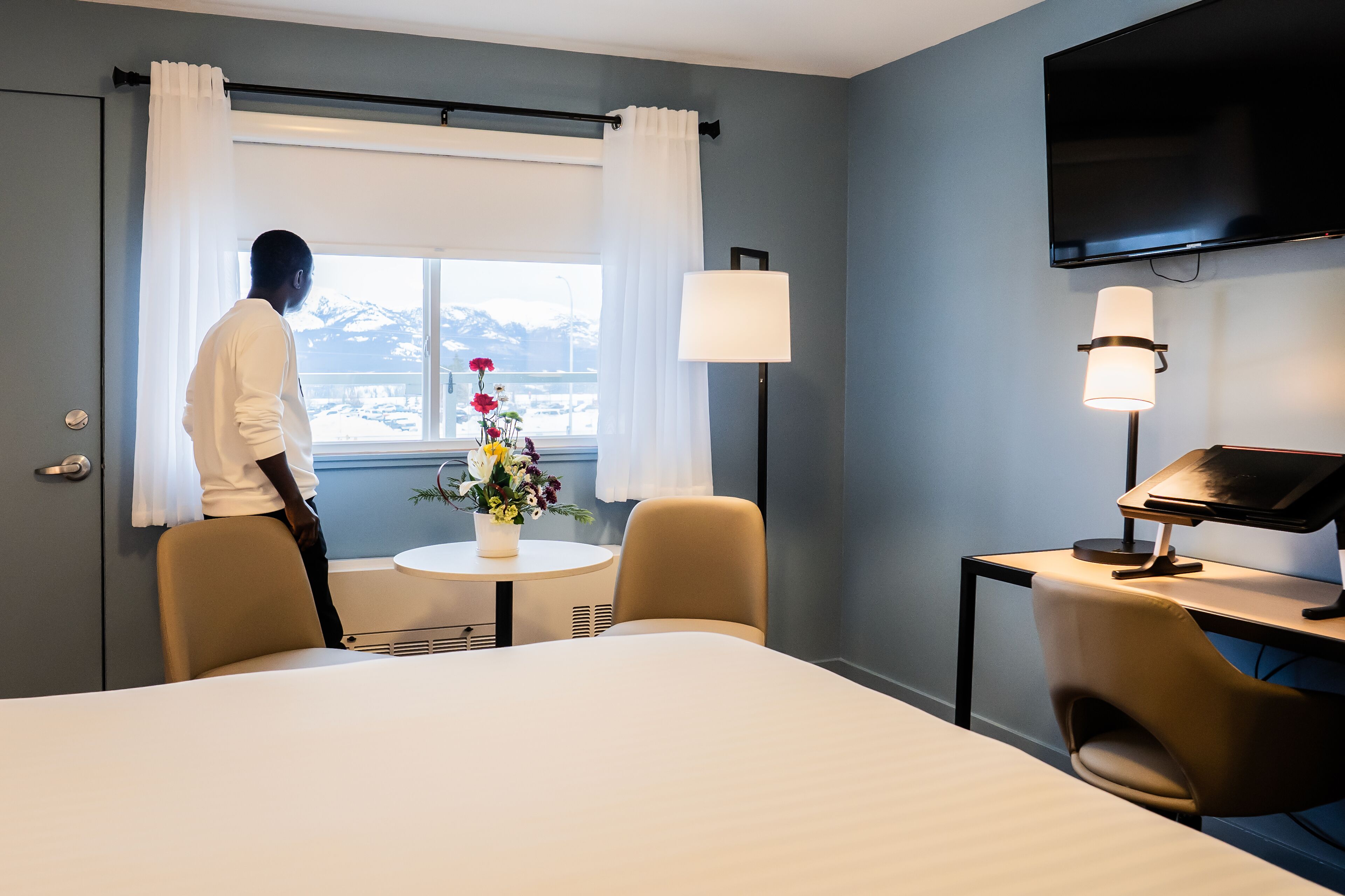 A hotel room with a bed, a desk with a computer, a chair, a small table with a vase of flowers, and a window with curtains.