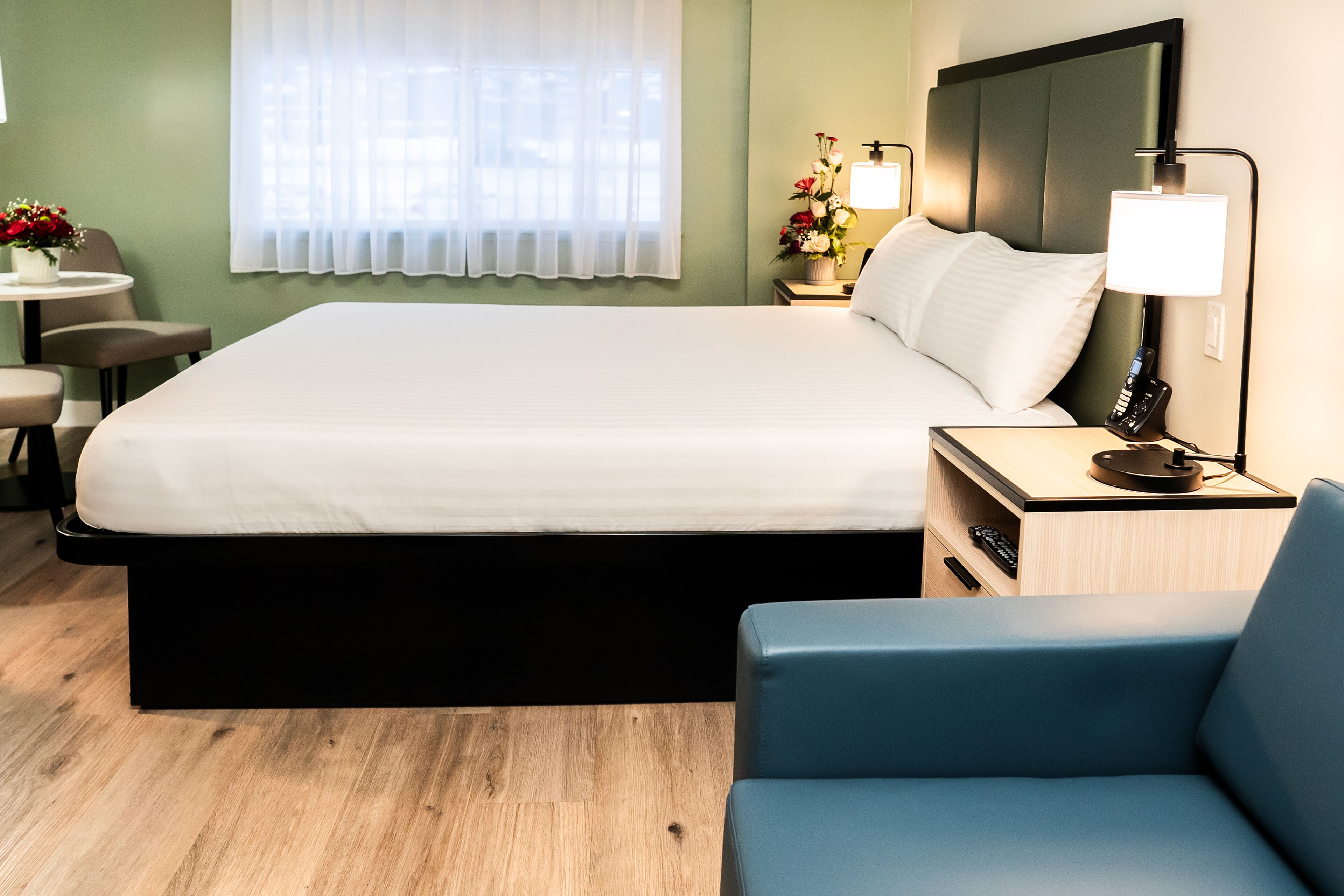 A hotel room with a large bed, a bedside table with a lamp, a chair, and a small table with flowers.