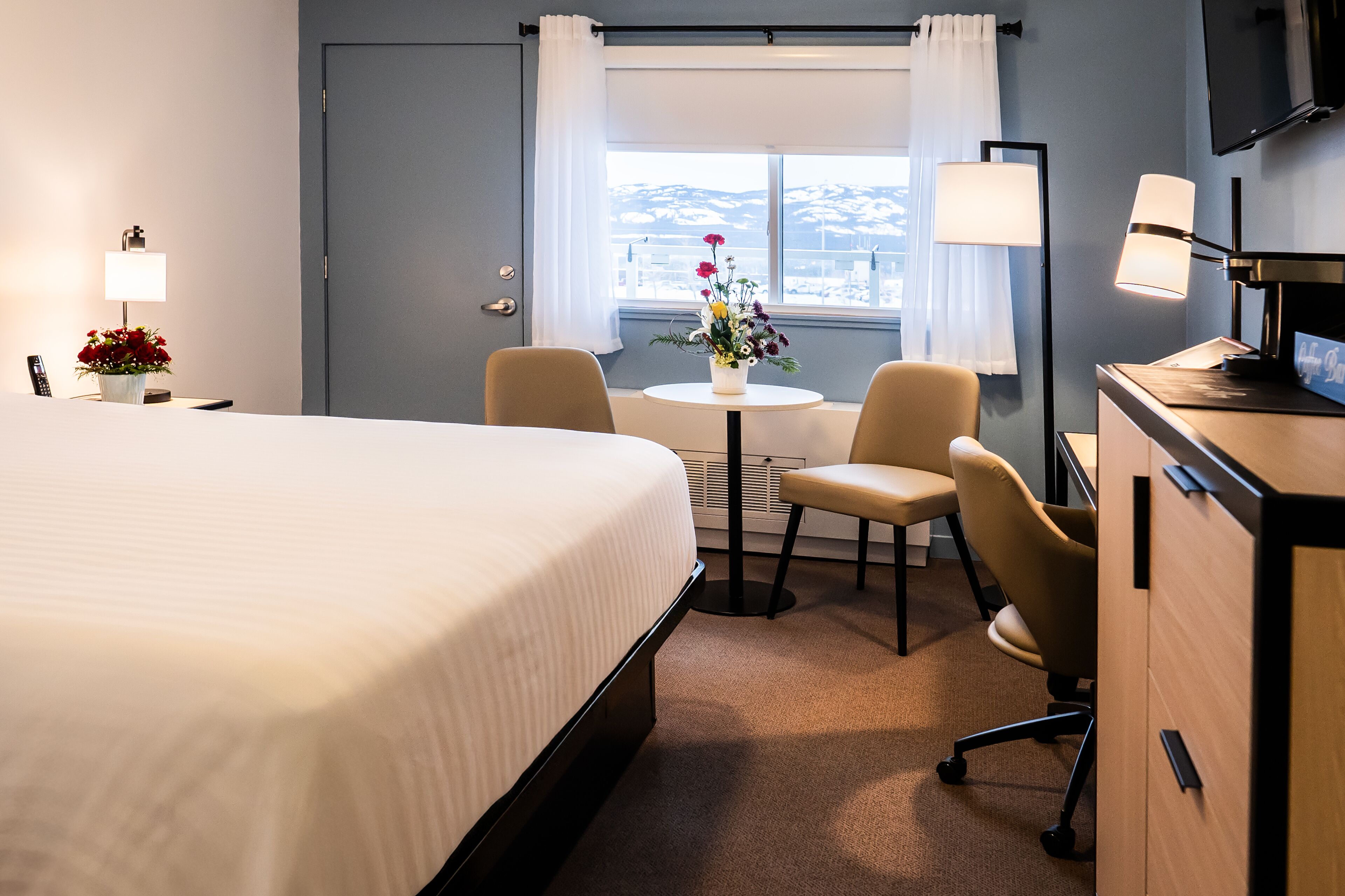 Habitación de hotel con una cama grande, un escritorio con computadora, una silla, una mesita con un jarrón de flores y una ventana con vista a la ciudad.
