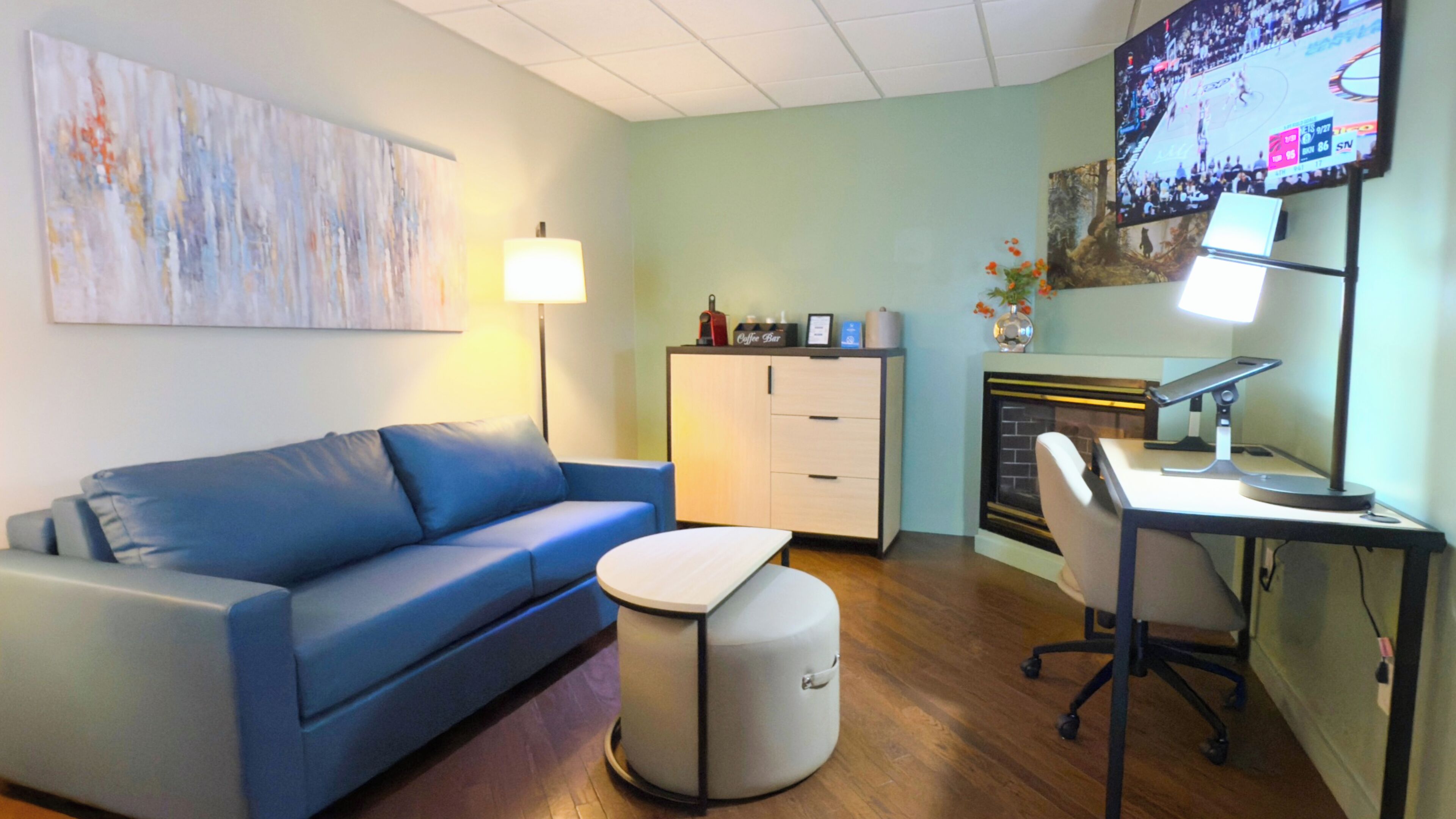 A room with a blue sofa, a small ottoman, a desk with a chair, a coffee machine, a fireplace, and a TV displaying a basketball game.