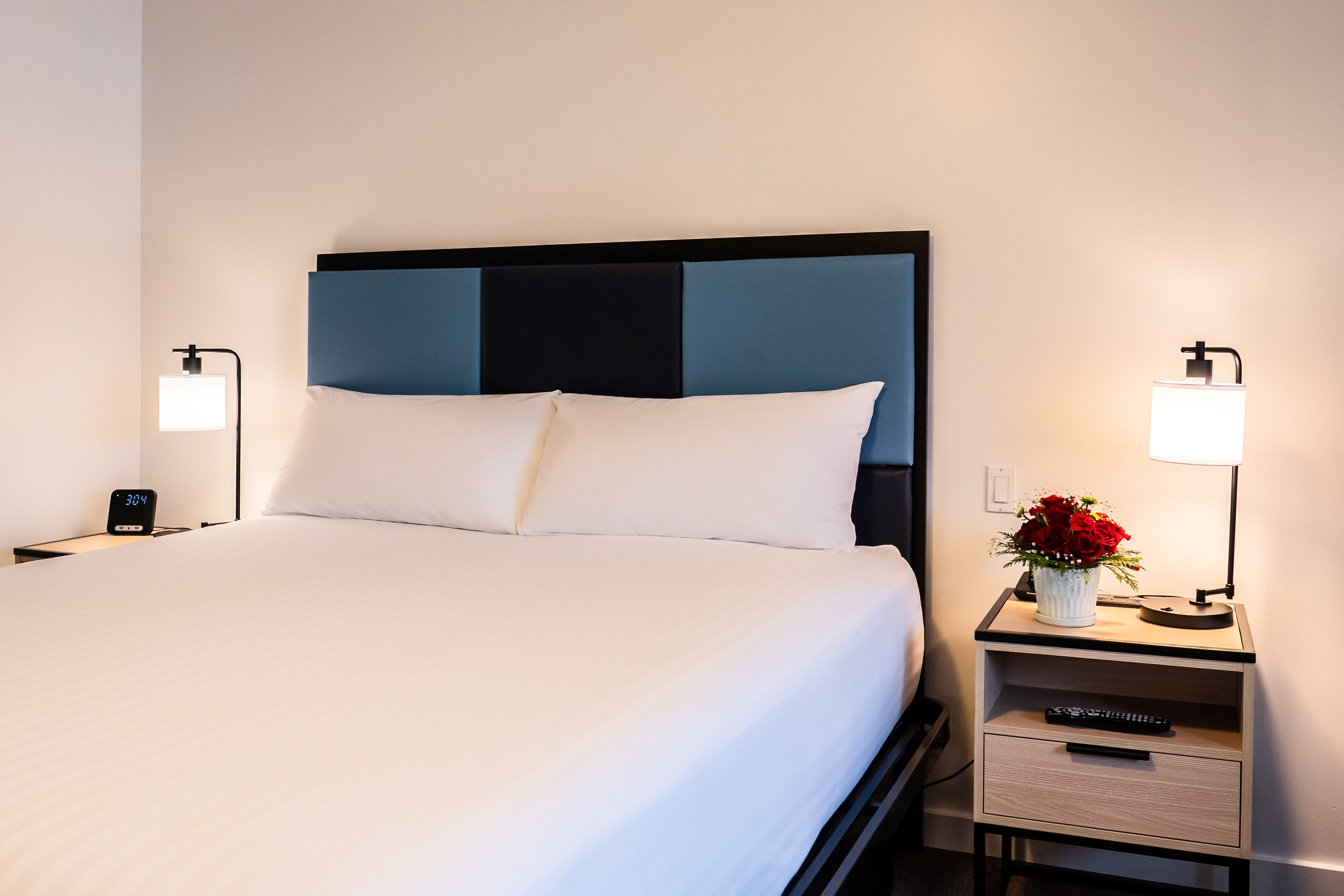 Ein ordentlich bezogenes Bett mit weißen Leinentüchern, ein moderner Kopfteil, Nachttischlampen, ein kleiner Tisch mit Blumen in einer Vase und ein Nachttisch mit Uhr und Fernbedienung.