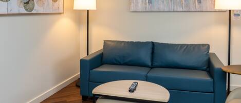 Un canapé bleu, une télécommande posée sur une petite table, une lampe et un tableau accroché au mur.