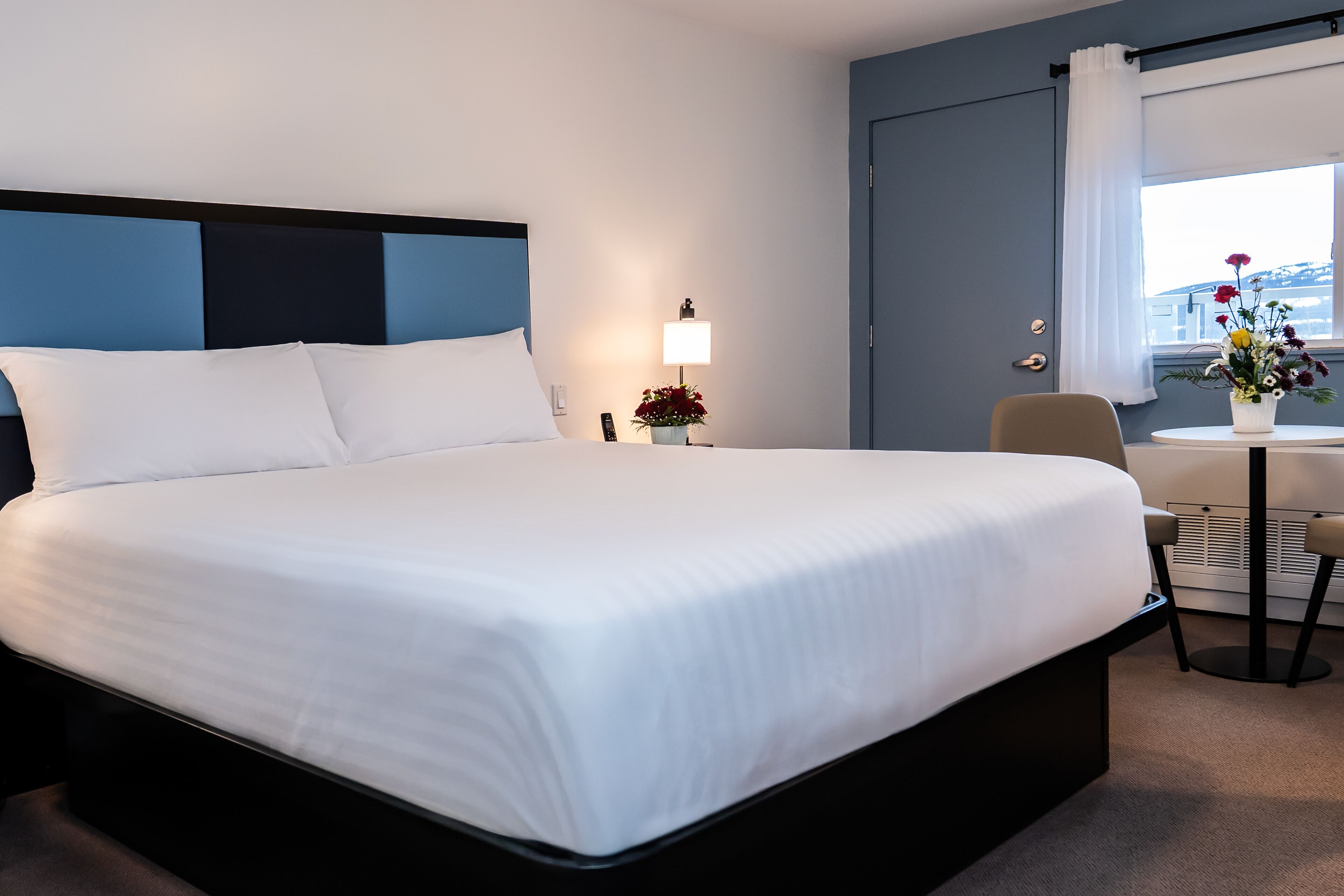 Habitación de hotel con una cama grande, una mesita de noche con lámpara, una silla y una pequeña mesa con un jarrón de flores.