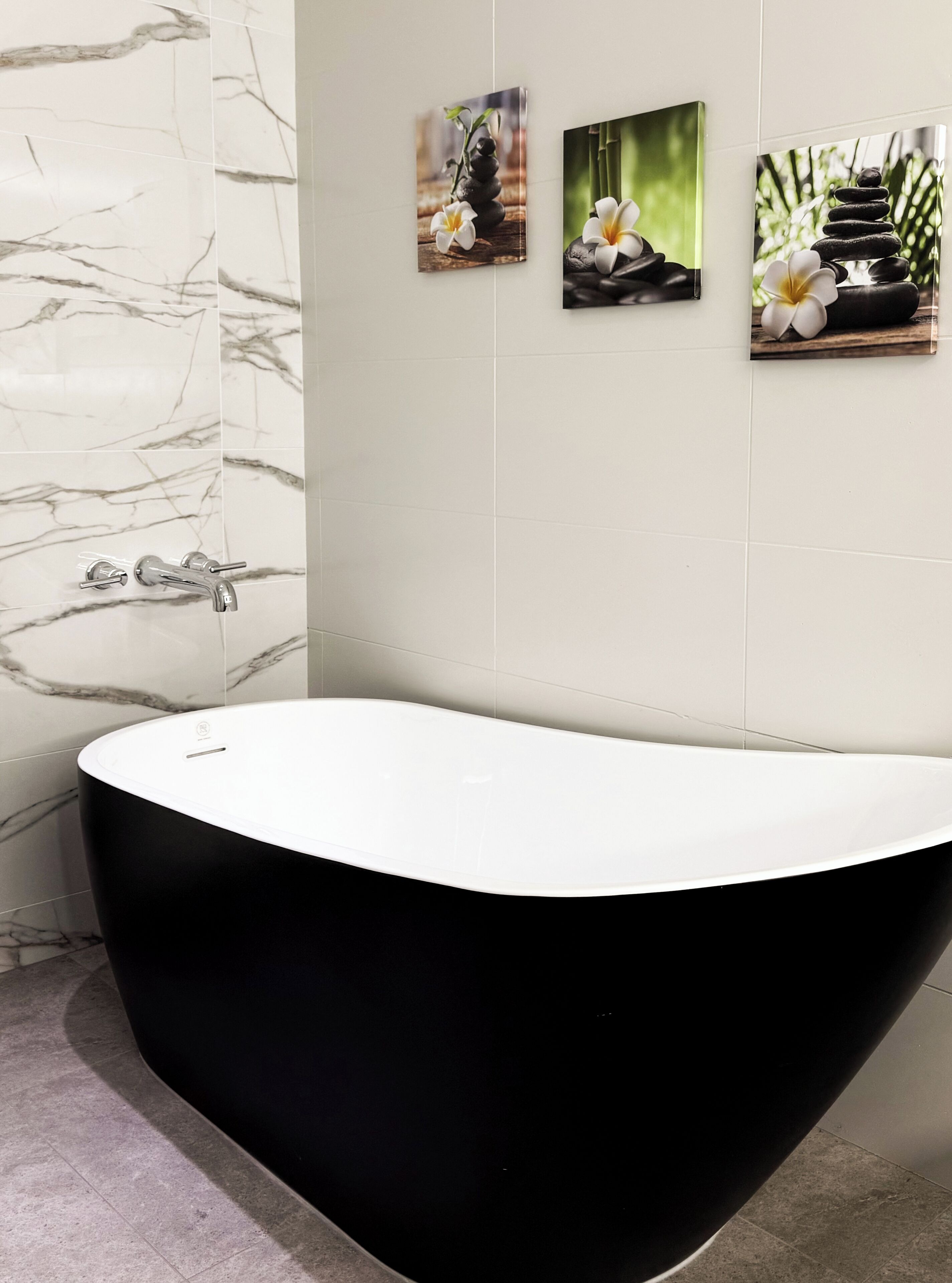 A modern bathroom with a freestanding black bathtub, marble wall tiles, and three framed pictures on the wall.