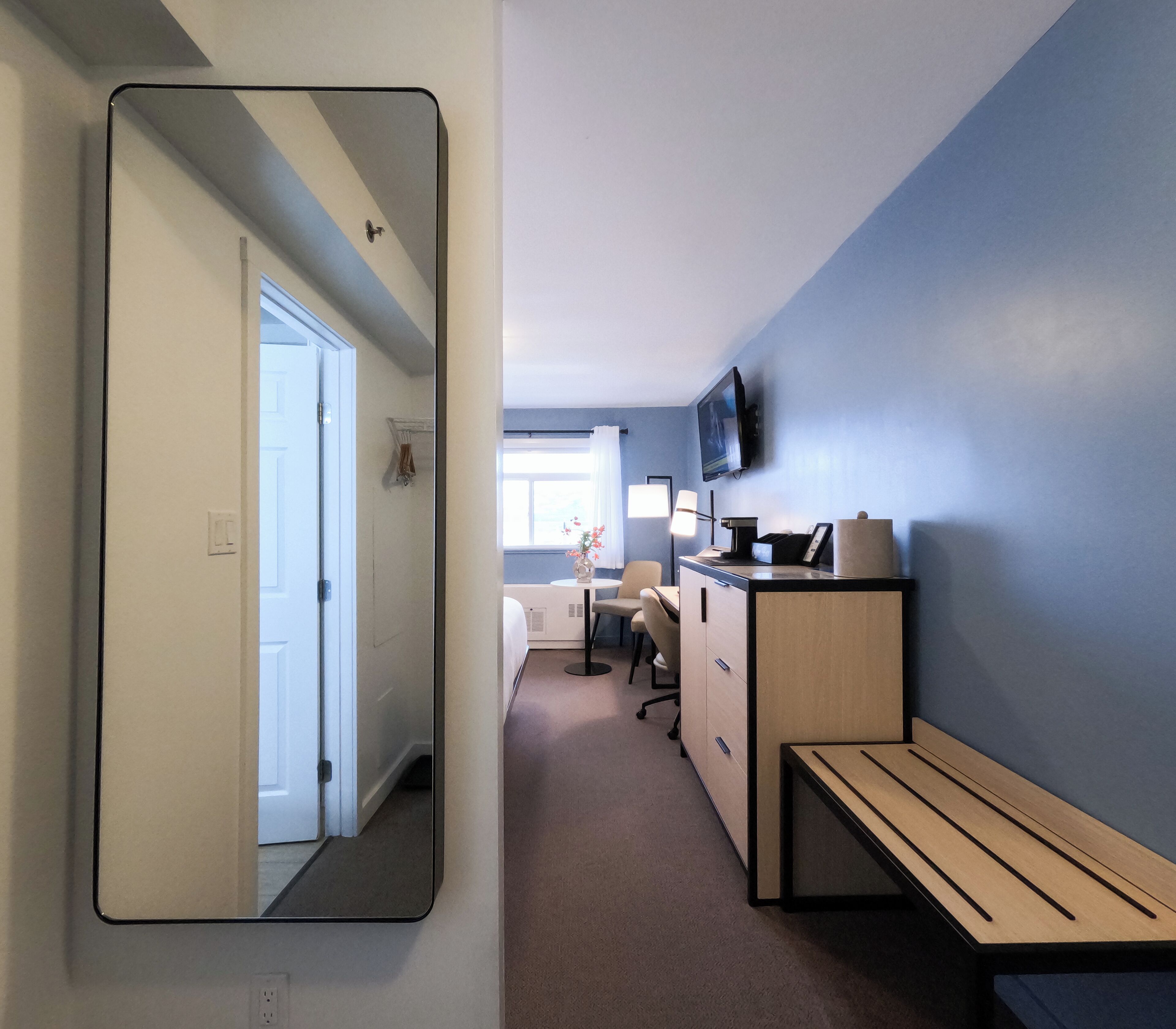 A hotel room with a large mirror, a desk with a chair, a bed, a TV mounted on the wall, and a window with curtains.