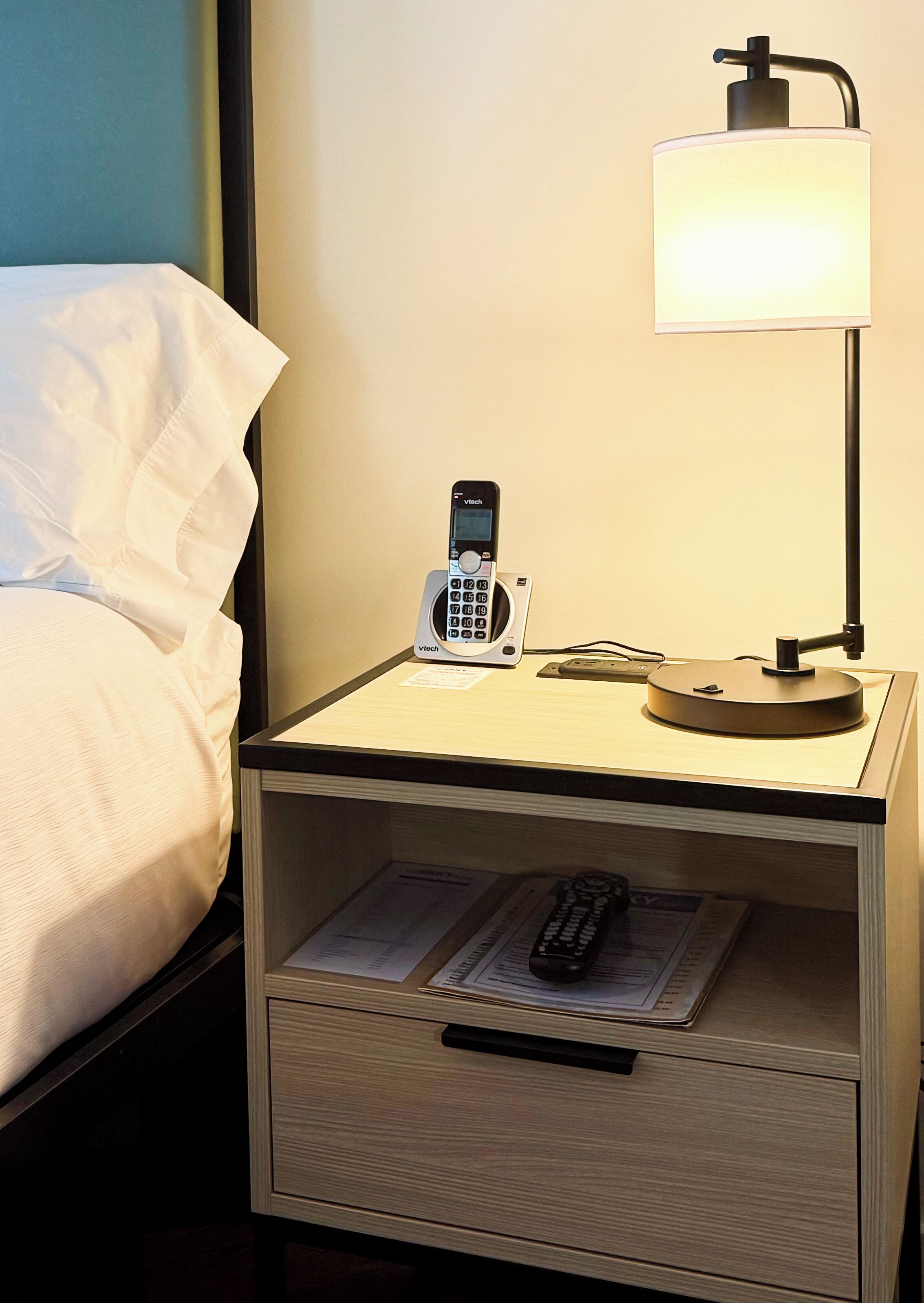 Een hotelkamer met nachtkastje, telefoon, lamp en een lade met een afstandsbediening.