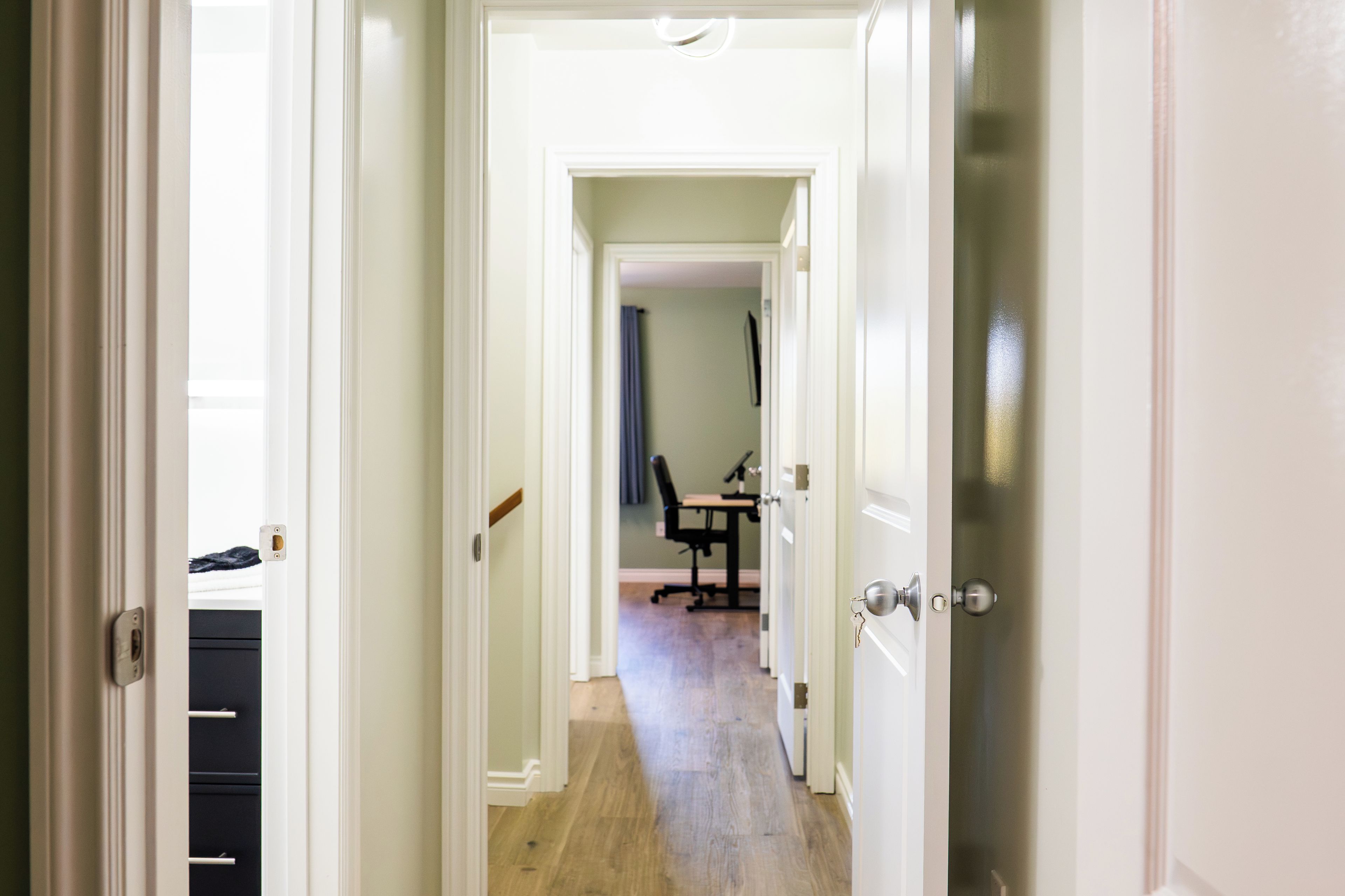 A hallway leading to a room with a desk and chair, a door with a key in the lock, and a cabinet with drawers.