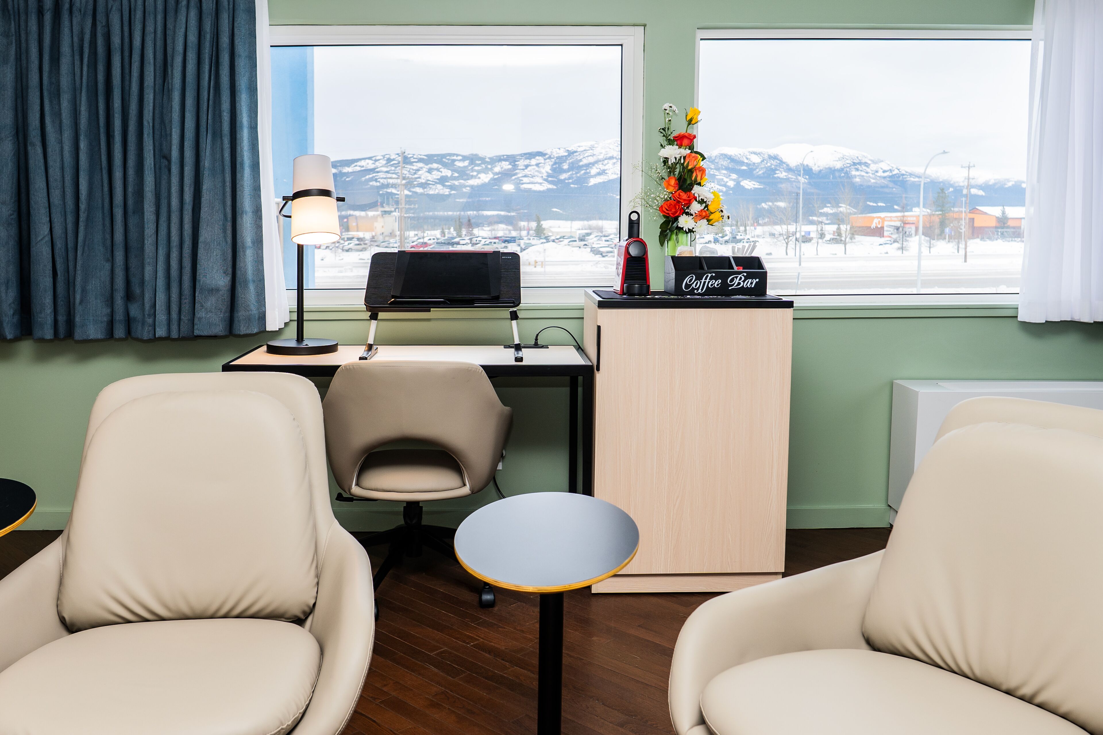 Une chambre d’hôtel équipée d’un bureau, de deux chaises, d’une lampe, d’un coin café et offrant une vue sur des montagnes enneigées.