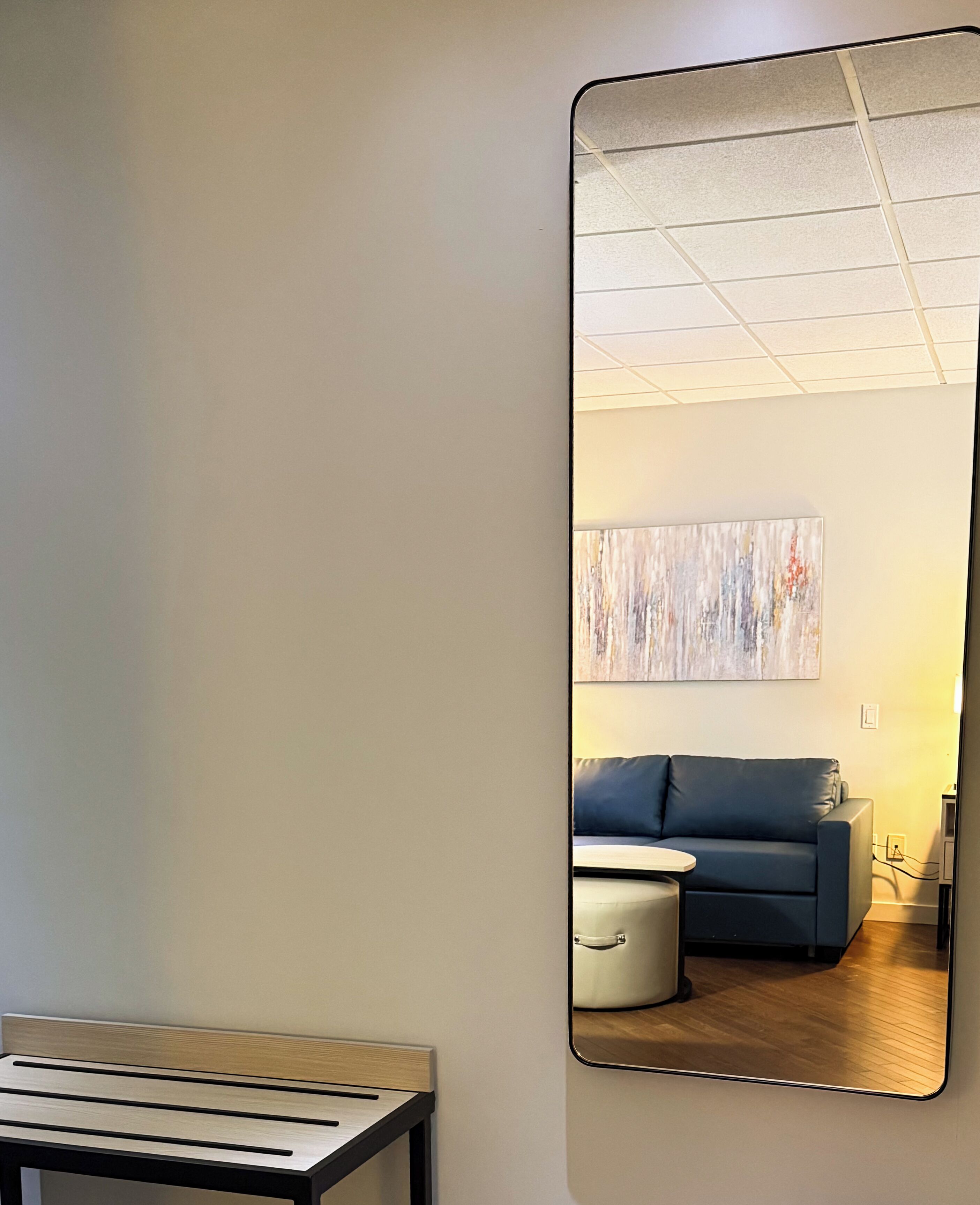 A room with a large mirror reflecting a blue sofa, a small table, and a painting on the wall.