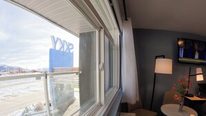 Habitación de hotel con un ventanal grande, vistas al exterior, un televisor en la pared y una pequeña mesa con un jarrón de flores.
