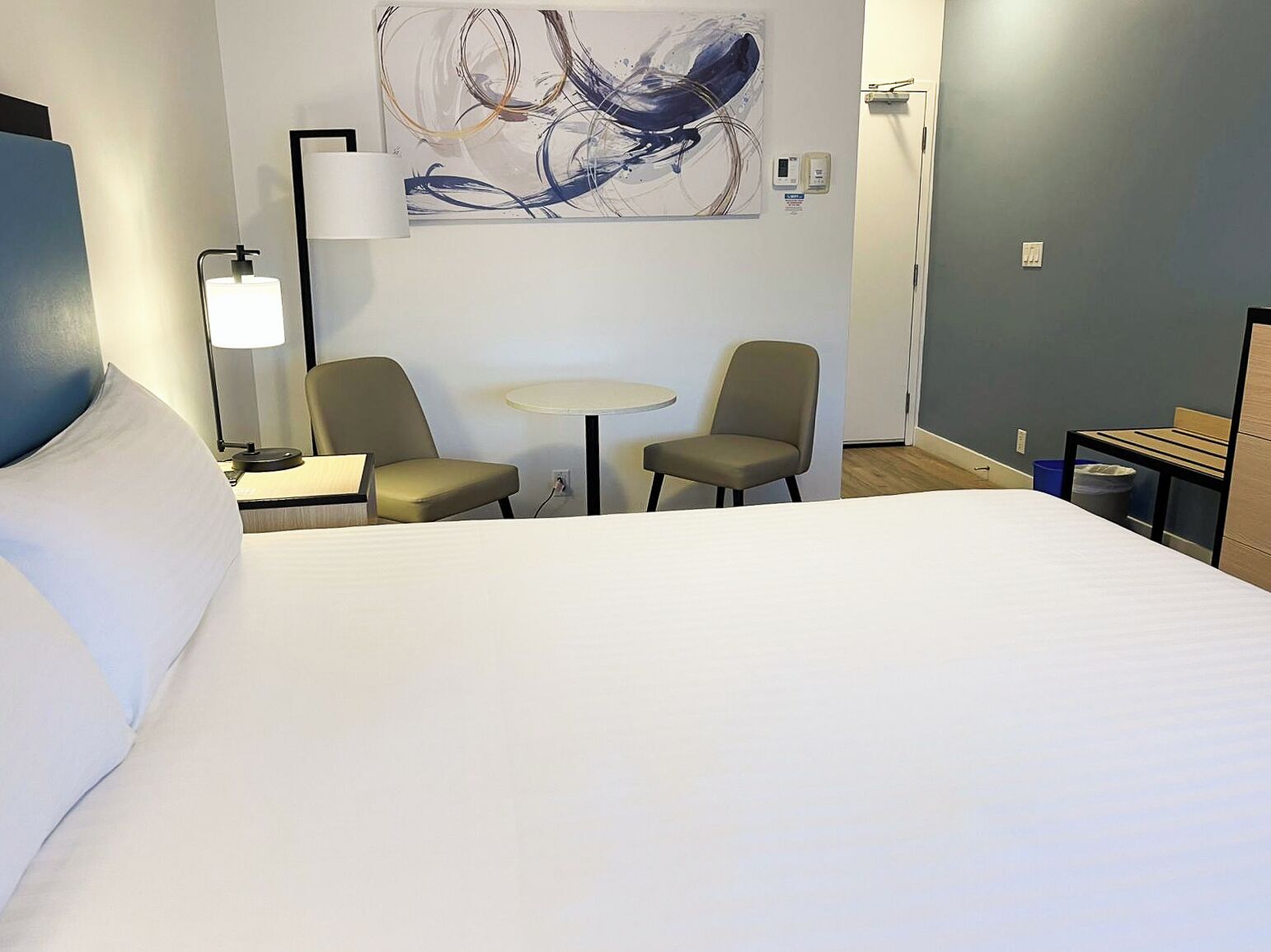 Ein Hotelzimmer mit einem großen Bett, zwei Sesseln, einem kleinen runden Tisch, einem Wandbild und einem Nachttisch mit Lampe.