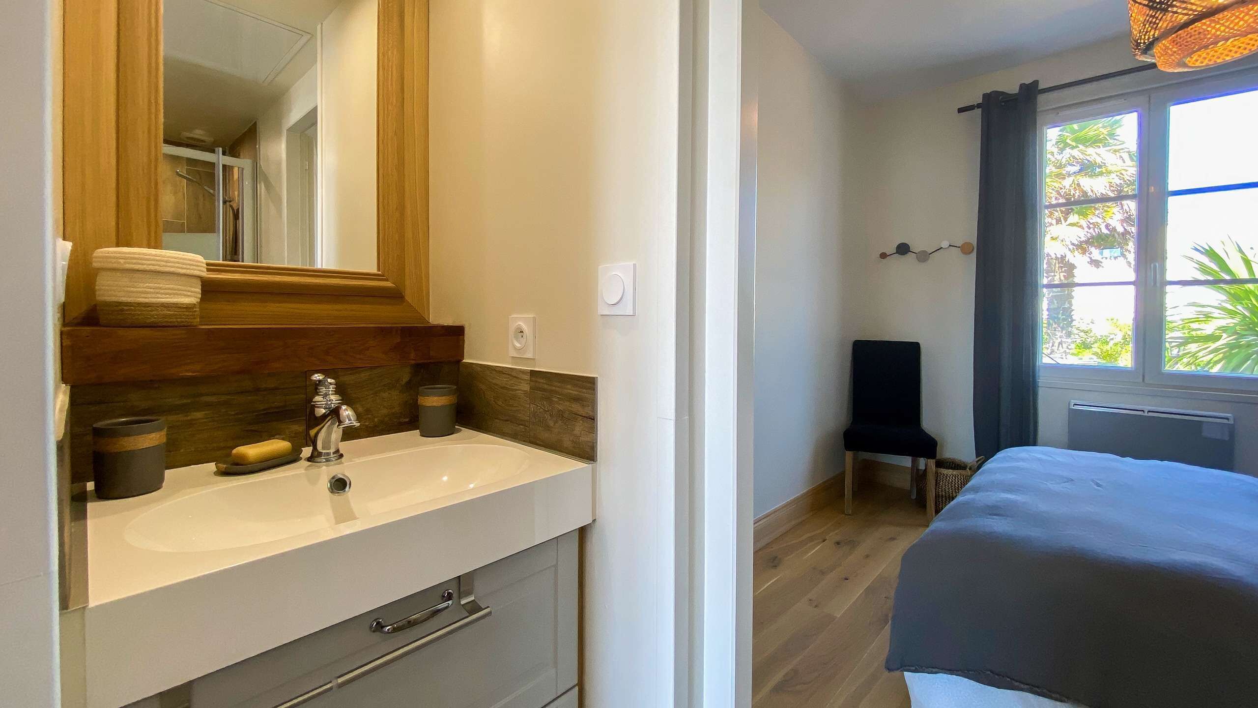 A hotel room with a bathroom featuring a sink, mirror, and towel storage. There is a window with a view of greenery outside.