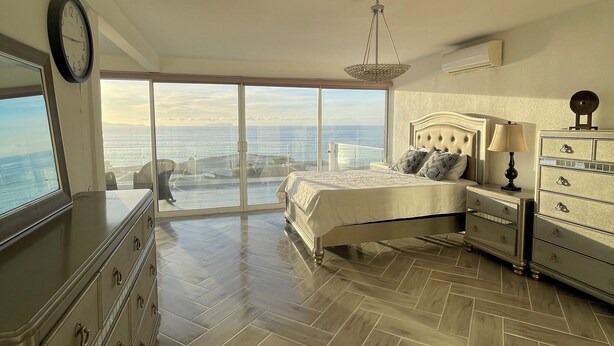 A bedroom with a large bed, a dresser, and a nightstand. There is a view of the ocean through large windows. - Beautiful 4 story beach house with amazing rooftop located in  Tijuana-Rosarito. (Tijuana)