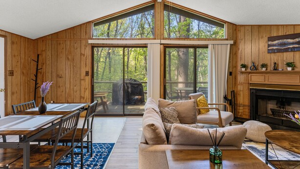 A cozy living room with a fireplace, a dining area with a table and chairs, and a large window overlooking a forest.