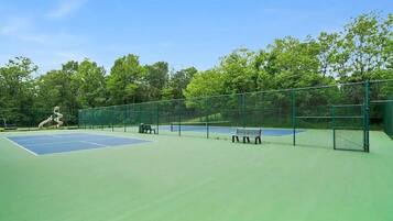 An outdoor tennis court with green fencing, two benches, and trees in the background.