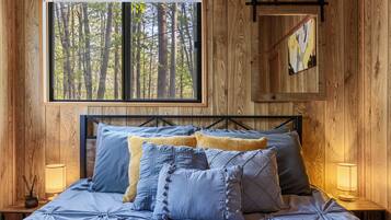 Un dormitorio con pared de madera, un ventanal con vista a un bosque, cama con ropa de cama azul y gris, y dos lámparas de noche.