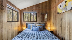 A wooden-paneled bedroom with a large bed, two windows, and a painting on the wall.