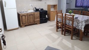Une cuisine compacte équipée d’un réfrigérateur, d’une cuisinière et de meubles de rangement en bois. On y trouve un coin repas avec une table et des chaises, ainsi qu’une petite fenêtre agrémentée de rideaux.